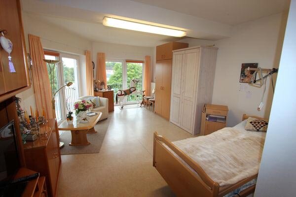 Ein heller, geräumiger Raum mit einem Bett, einem Tisch und Stühlen. Auf dem Tisch steht eine Blumenvase. Große Fenster bieten einen Blick nach draußen. Ein Kleiderschrank und weitere Möbel ergänzen den Raum. Die Atmosphäre wirkt freundlich und einladend.