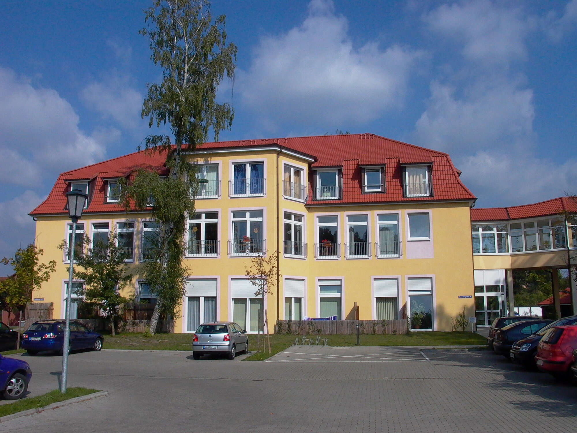Ein gelbes Gebäude mit rotem Dach, das mehrere Fenster aufweist. Umgeben von einem Parkplatz mit Fahrzeugen. Im Hintergrund sind Bäume und ein blauer Himmel zu sehen.