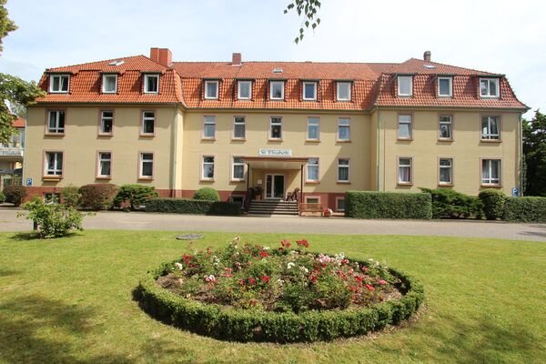 Ein mehrstöckiges, gelbes Gebäude mit roten Dachziegeln. Es gibt viele Fenster und einen Eingangsbereich mit Treppen. Vor dem Gebäude befindet sich ein gepflegter Rasen mit einem runden Blumenbeet in der Mitte.