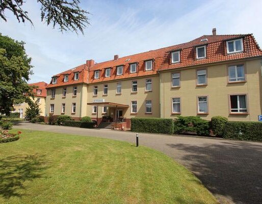 Ein mehrstöckiges Gebäude mit gelber Fassade und roten Dachziegeln. Es verfügt über mehrere Fenster und einen Eingang mit Treppe. Vor dem Gebäude erstreckt sich eine grüne Rasenfläche, umgeben von Bäumen und einem kleinen Parkplatz.