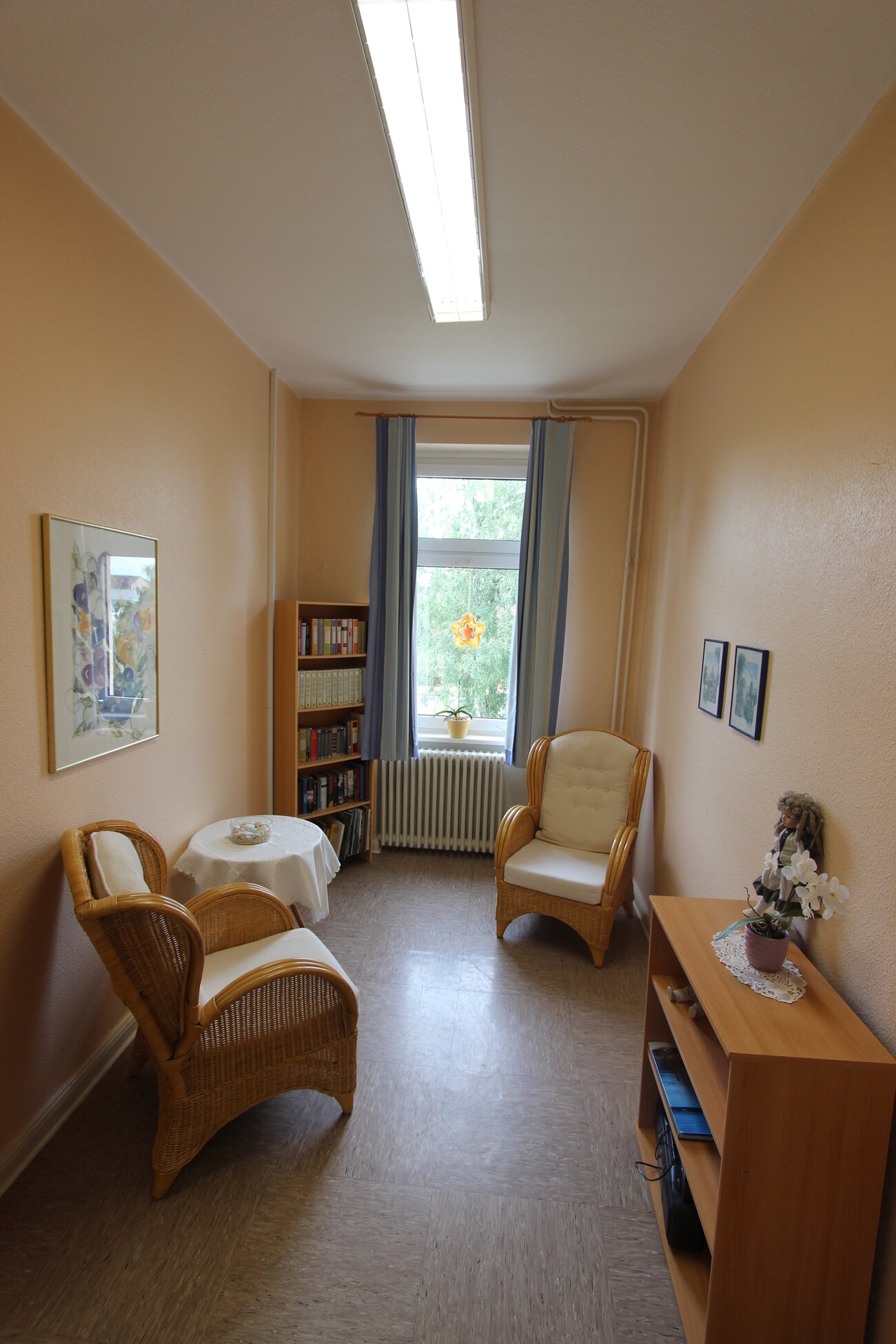 Ein schmaler Raum mit zwei Korbstühlen, einem kleinen Tisch und einem Bücherregal. Ein Fenster mit Vorhängen lässt Tageslicht herein. An einer Wand hängen zwei Bilder und auf dem Tisch steht eine dekorative Vase.