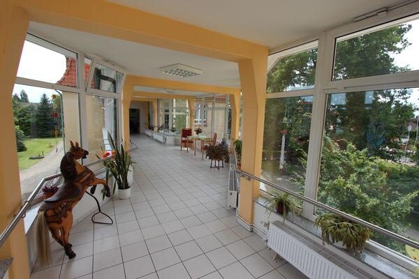 Ein heller Flur mit großen Fenstern, der von Pflanzen gesäumt ist. Auf der linken Seite steht eine Holzskulptur eines Pferdes. Am Ende des Flurs sind einige Möbel und ein Blick auf den Außenbereich mit Bäumen und Grünflächen zu sehen.