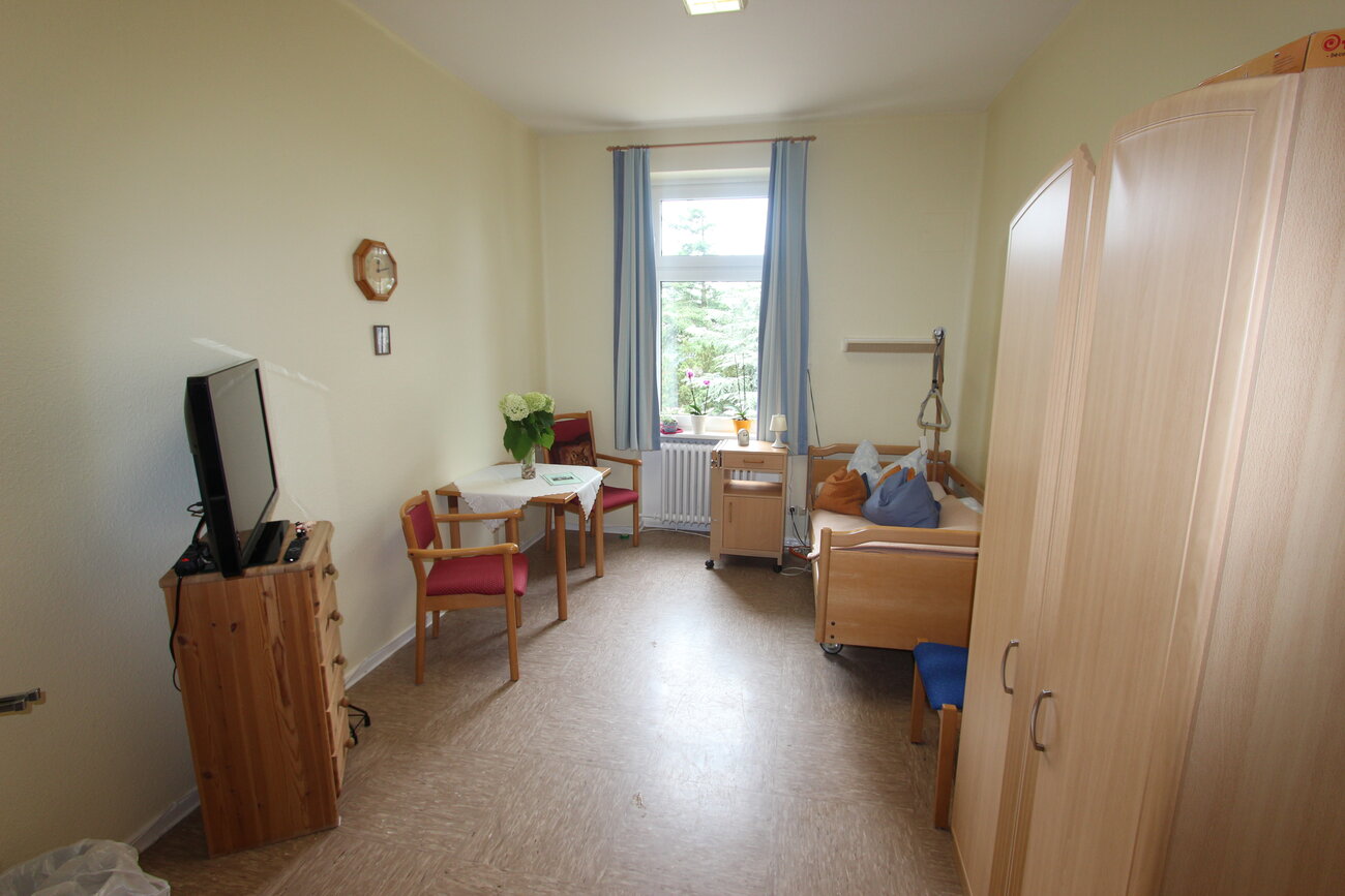 Ein heller, geräumiger Raum mit einem Bett, einem Tisch und Stühlen. An den Wänden hängen ein Uhr und ein Fenster mit Vorhängen. Eine Kommode und ein Fernseher stehen zur Seite. Der Raum wirkt einladend und freundlich.