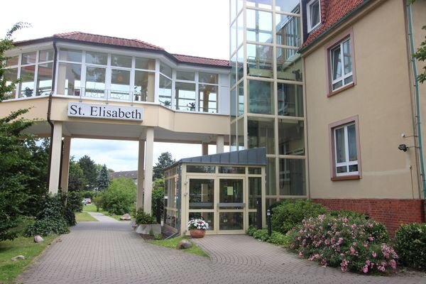 Die Aufnahme zeigt den Eingangsbereich des Gebäudes "St. Elisabeth". Es ist ein mehrstöckiges Haus mit großen Fenstern und einem Aufzug. Der Eingangsbereich ist von Pflanzen und Wegen umgeben, die zum Parkplatz führen.