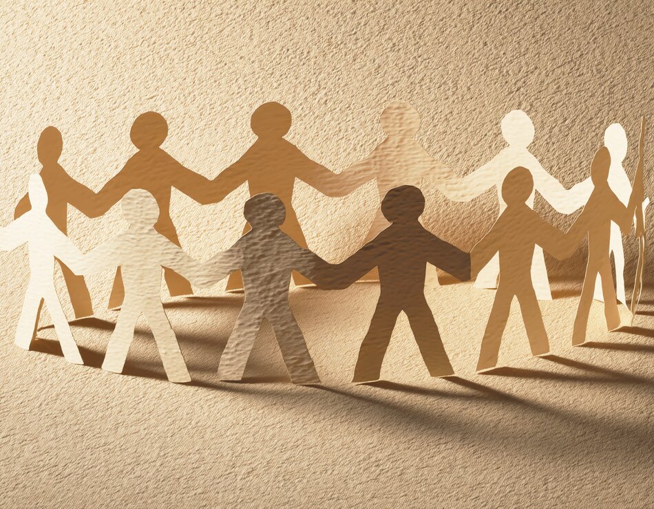 Eine Gruppe von aus Papier ausgeschnittenen Figuren steht Hand in Hand und bildet einen Kreis. Die Figuren sind in verschiedenen Tönen gehalten, was die Vielfalt und den Zusammenhalt symbolisiert. Der Schattenwurf erzeugt eine dreidimensionale Wirkung auf dem Untergrund.