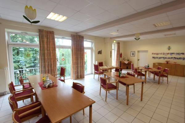 Ein heller Raum mit mehreren Tischen und Stühlen. Große Fenster lassen Tageslicht herein. Die Wände sind mit Bildern dekoriert. Einige Tische sind für Gäste gedeckt, was eine einladende Atmosphäre schafft. Grünpflanzen sind sichtbar, was zur freundlichen Umgebung beiträgt.